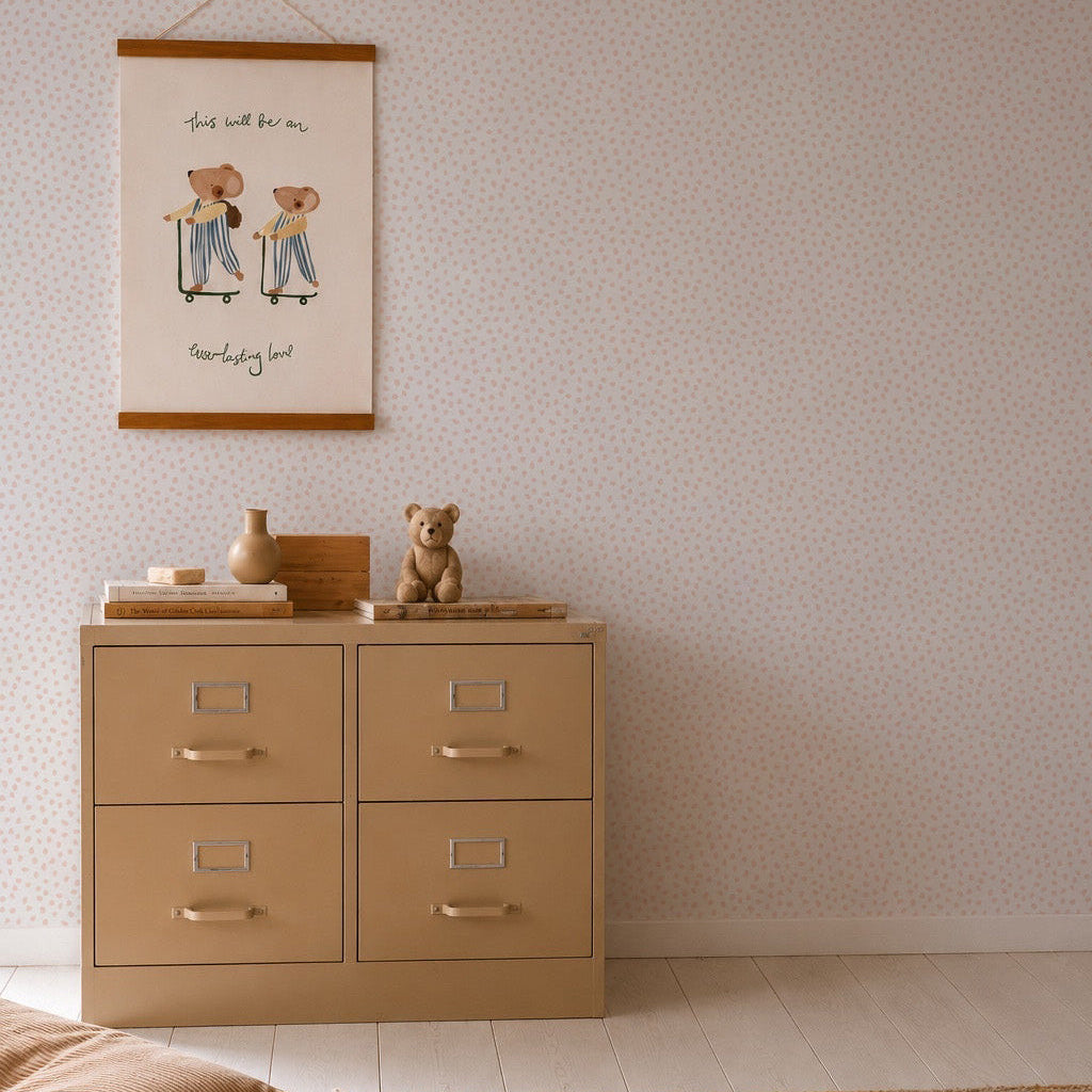 A beige dresser with four drawers stands against a wall decorated in Sprinkle Wallpaper Sample, Pink. On top, a teddy bear, stacked books, and small decor are displayed. Above hangs a framed print featuring two figures and handwritten text.