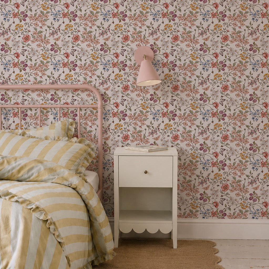 A cozy bedroom features Sunlit Butterfly Meadow Wallpaper, a pink metal bed with yellow and white striped bedding, a small white nightstand, a pink wall lamp, and a tan woven rug on a white wooden floor.