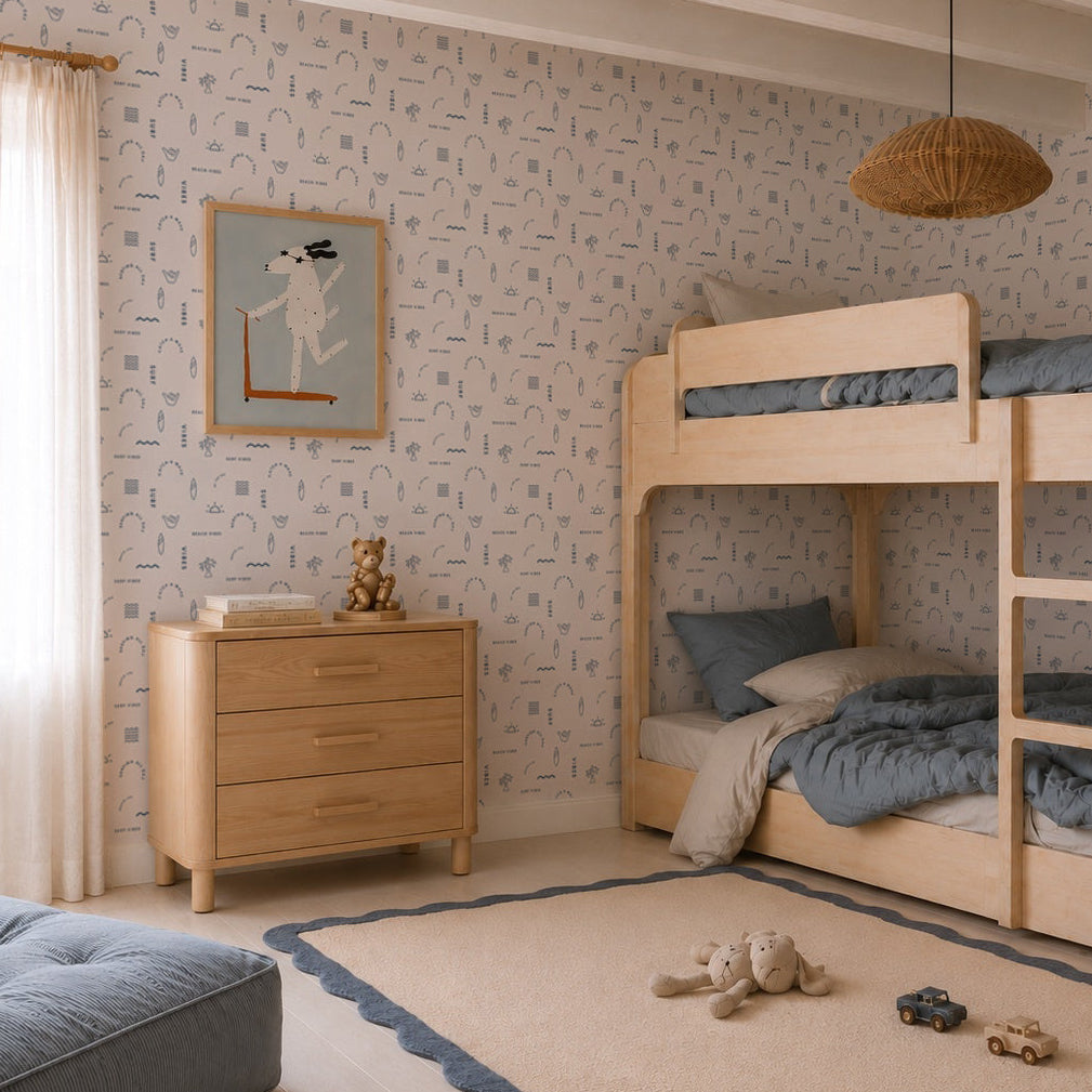 A cozy children’s room features light wood bunk beds, blue bedding, a matching dresser, a beige rug with blue trim and toys, plus Beach Vibes Wallpaper in Blue; sunlight pours in through a window with sheer curtains.