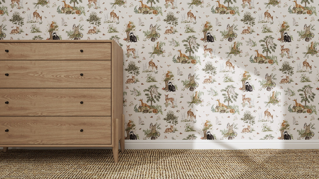 A wooden dresser stands on a woven carpet beside a wall covered in Tales Beneath The Trees Wallpaper Cream, featuring playful giraffes, lions, monkeys, and trees. Sunlight highlights the vibrant colours of this high-quality animal-themed wallpaper.