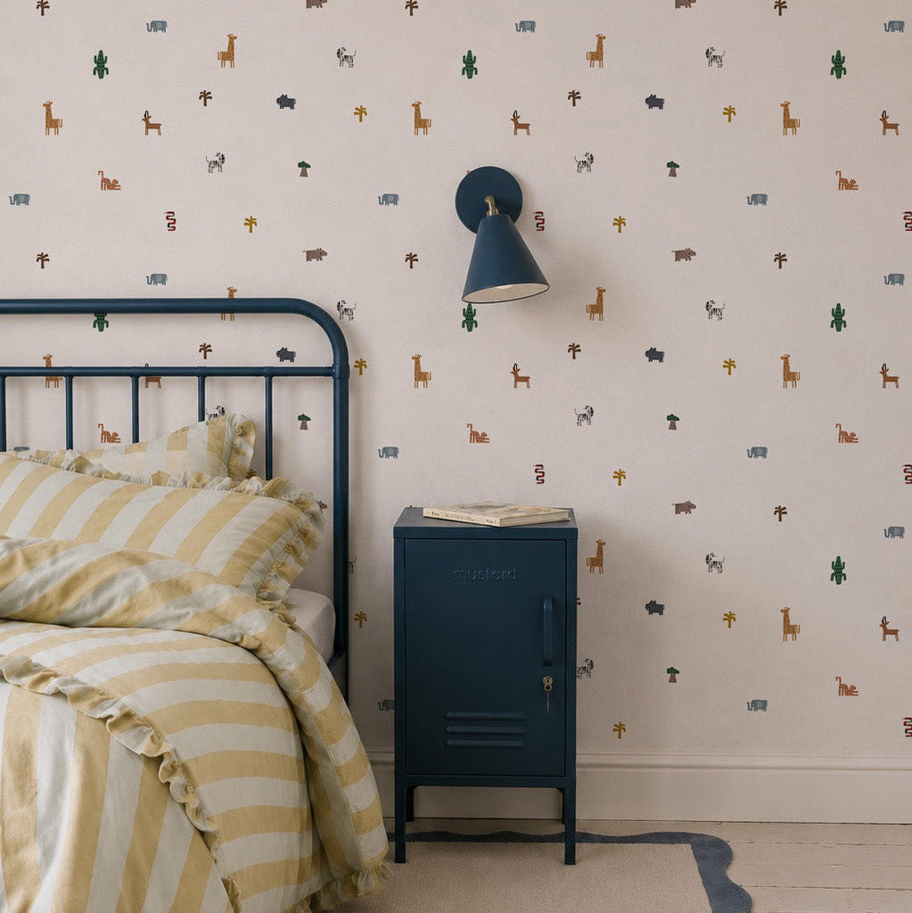 A cozy bedroom corner with a metal bed frame, yellow striped bedding, blue nightstand, wall-mounted lamp, and playful Tiny Tracks Safari Wallpaper—soft animal prints that add a whimsical touch to any kid’s room.