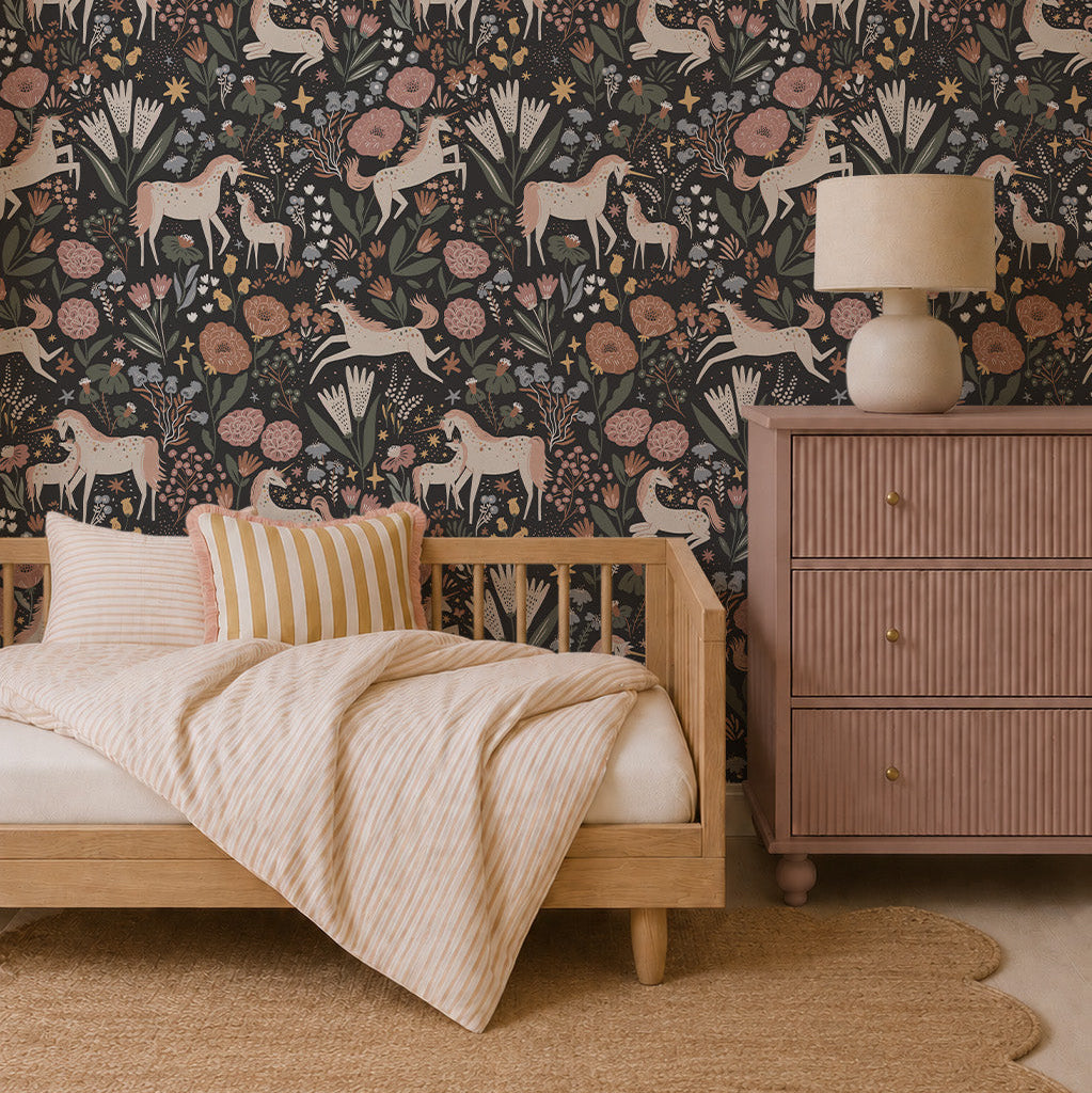 A cozy bedroom with a wooden daybed, striped bedding, round rug, and a pink dresser topped with a lamp. The wall features whimsical design touches with Unicorn Meadow Wallpaper in Charcoal, featuring muted floral and horse patterns.