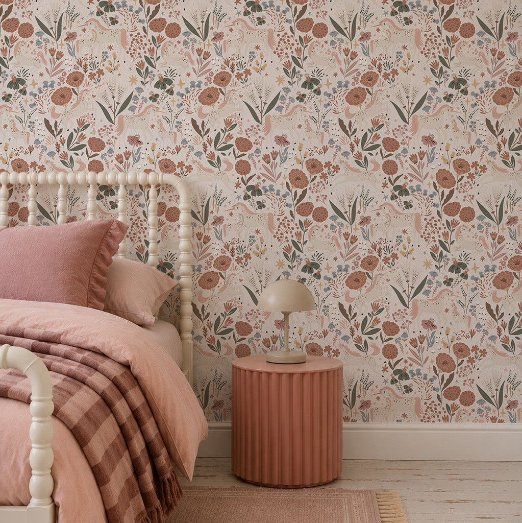 A cozy bedroom features a spindle bed with pink bedding, a plaid blanket, and a round ribbed nightstand topped with a beige lamp. The girly decor is enhanced by the Unicorn Meadow Wallpaper in cream.