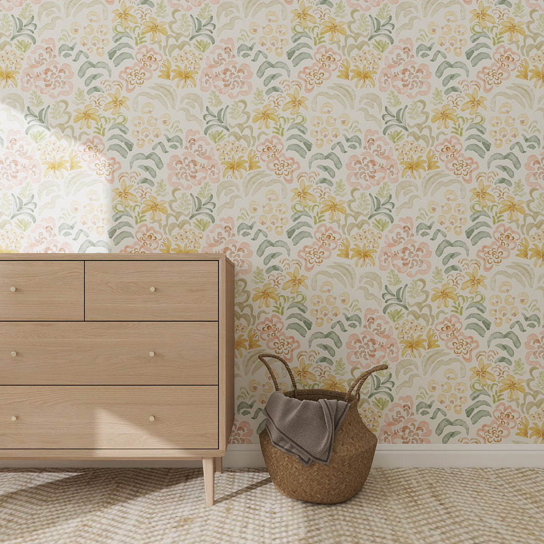 A light wooden dresser sits on a textured beige rug by a woven basket with a grey blanket, set against Floret Dreamscape Wallpaper in green, pink, and yellow floral patterns. Sunlight streams in from the left.