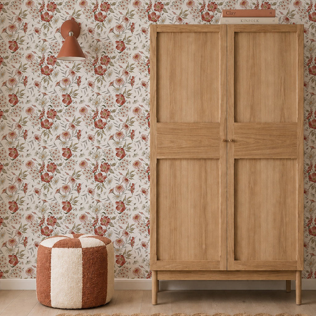 A wooden wardrobe stands next to a red and cream pouf on a tan woven rug, with Wild Rose Dreams Wallpaper, a wall lamp, and books stacked on top of the wardrobe.