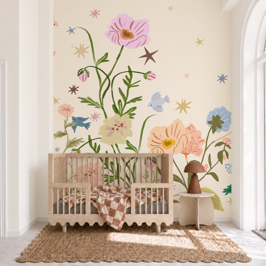 A wooden crib, The Wishing Garden, Cream, sits in front of a wall featuring a whimsical pink floral wall mural decorated with stars and small birds.