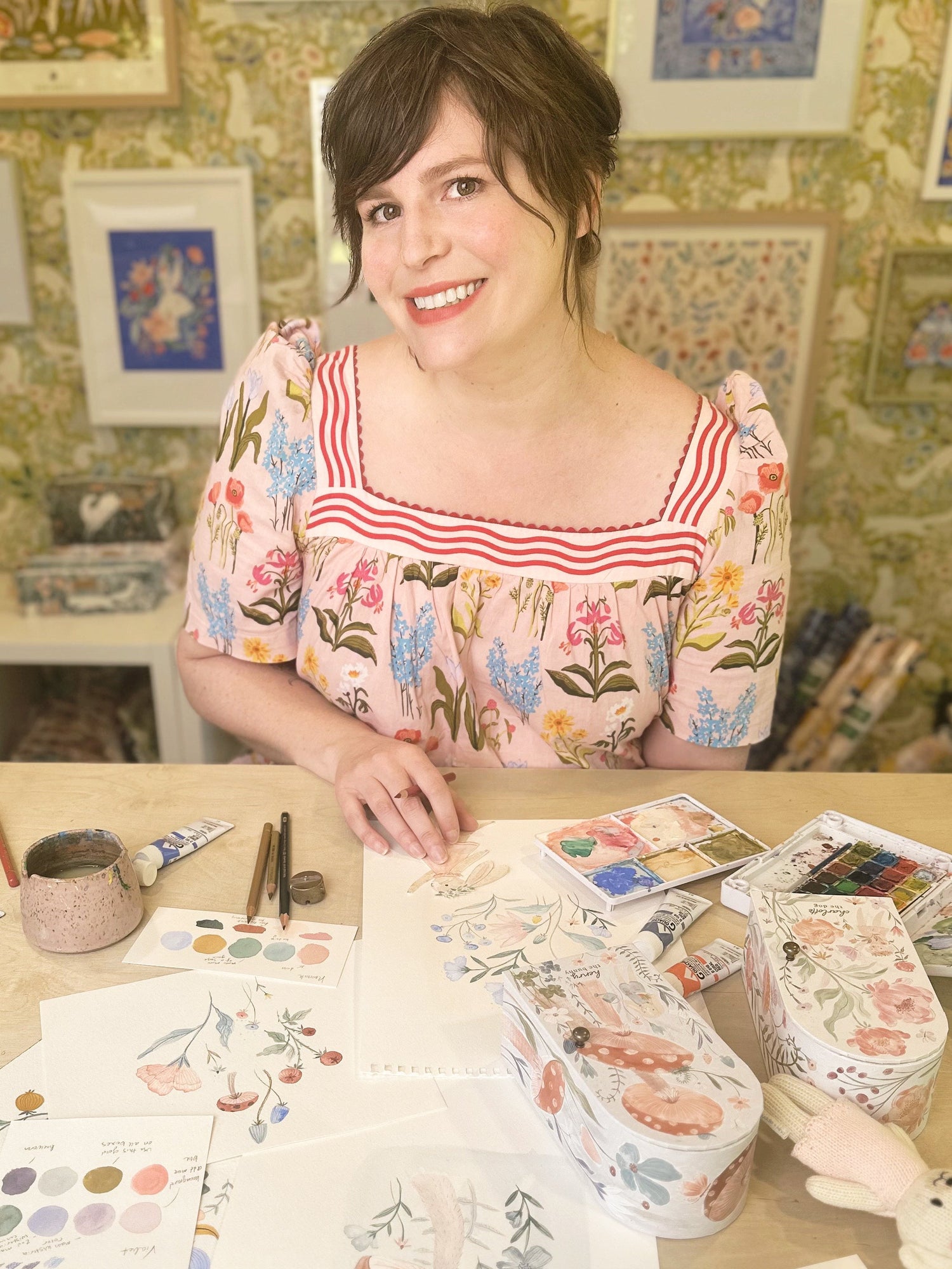 A woman in a floral dress smiles at a table covered with watercolor paintings, paintbrushes, and art supplies, with colorful floral artwork displayed on the wall behind her.
