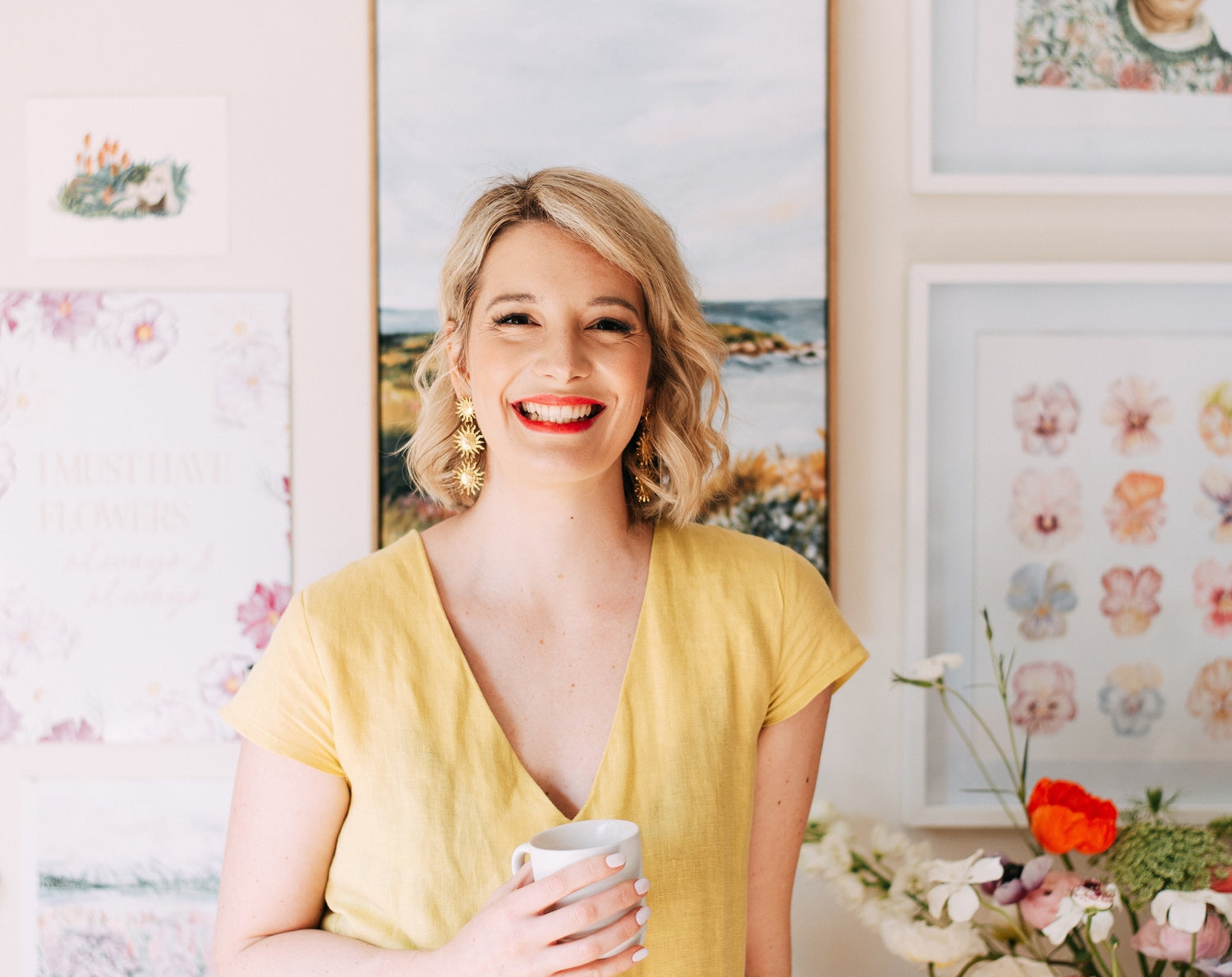 A smiling woman in a yellow dress holds a mug, standing in front of a wall decorated with colorful art and flowers. She has short blonde hair and bright red lipstick.
