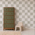 A green and wood dresser stands next to a small beige chair, with a checkered rug on the floor and stylish Billys Checkered Wallpaper in Sage as the background, creating a charming wall decor accent for the room.