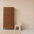 A brown wooden dresser stands next to a small beige chair on a light wood floor, with Butterfly Meadowland Wallpaper in Dune adding a touch of nature and a checkered rug completing the room.