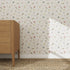 A mid-century modern wooden dresser stands on a woven rug near a wall featuring the Powdered Petals Wallpaper sample. Sunlight casts a shadow, creating a bright and airy atmosphere.