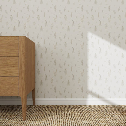 A wooden dresser with three drawers stands on a woven rug by a white wall decorated with Delicate Meadows Wallpaper | Sample, featuring a subtle beige floral pattern. Sunlight casts a diagonal shadow, enhancing the décor’s colour and quality.