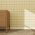 A wooden dresser with three drawers stands on a woven rug beside a yellow Checkered Wallpaper Repeat Pattern, highlighting the vibrant wallpaper color as sunlight casts a shadow from the right.