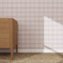 A wooden dresser with three drawers sits on a woven rug beside a wall covered in Checkered Wallpaper Repeat Pattern Pink | Sample. Sunlight casts a shadow, highlighting the quality of the wallpaper samples pink checks.