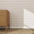 A wooden dresser stands on a woven rug against the Checkered Wallpaper Repeat Pattern Cream, its refined plaid design highlighted by sunlight casting geometric shadows and enhancing the room’s bright, minimalist appeal.