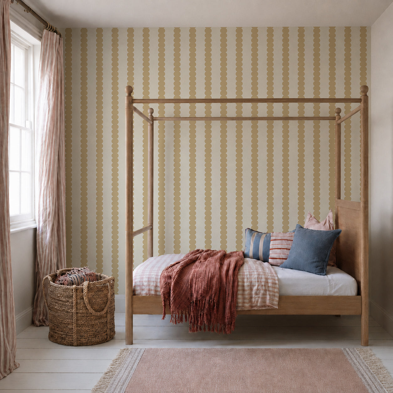 A cozy bedroom featuring a wooden canopy bed, walls accented with Scallop Stripe Wallpaper in Yellow, pink and blue pillows, a rust-colored throw, striped curtains, a large woven basket, and sunlight streaming through a window.