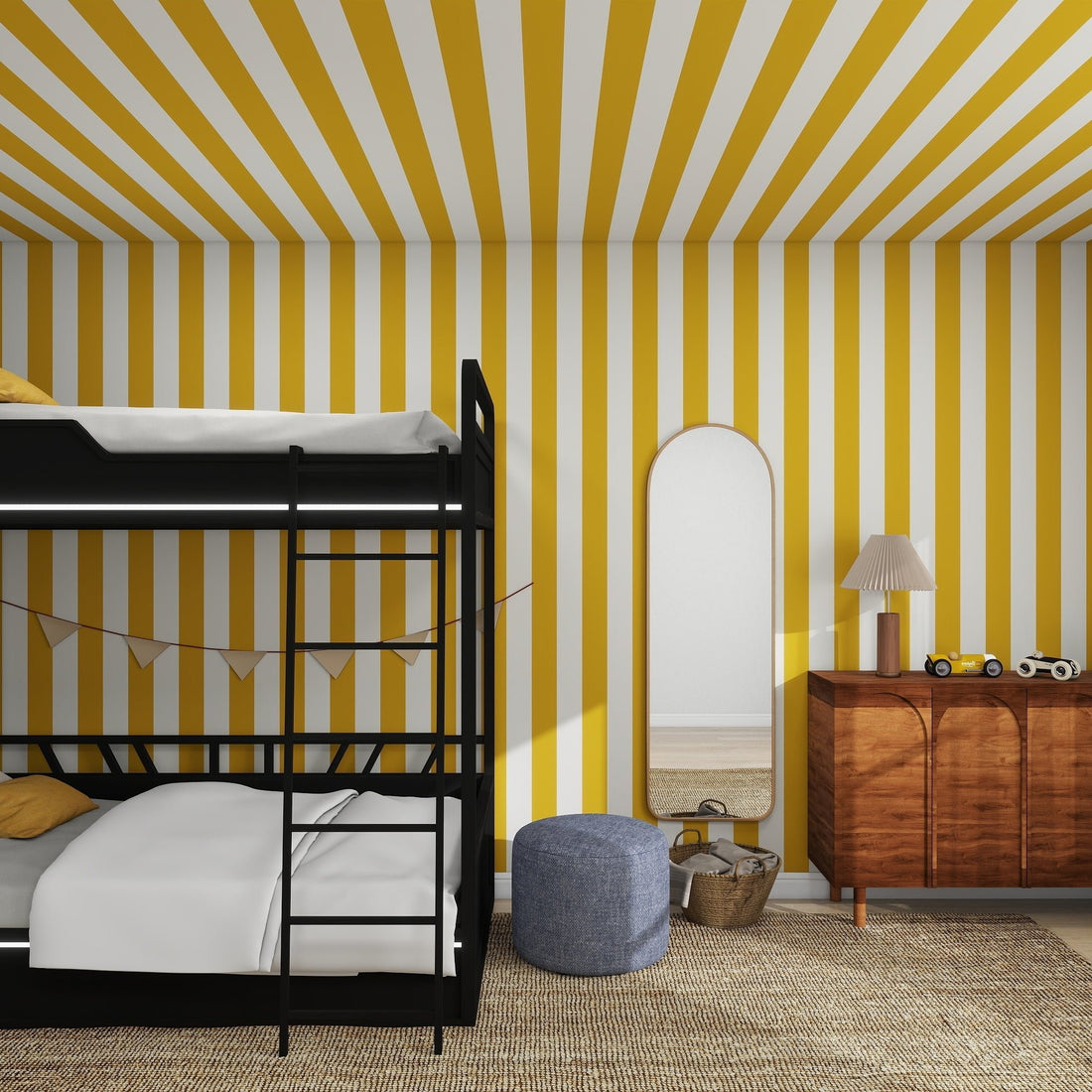 The childrens room showcases munks and mes Circus Stripe Wallpaper in mustard and white. A black bunk bed is on the left, while a wooden dresser with a lamp and toy cars sits under an oval mirror on the right. A gray pouf adds charm to this delightful decor.