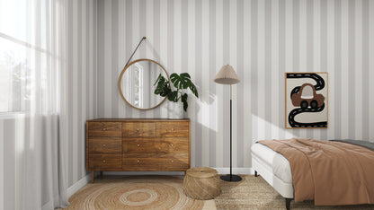 The bedroom, with Circus Stripe Wallpaper Stone by munks and me, showcases a wooden dresser topped with a plant, a round mirror, and a floor lamp. A bed with brown bedding sits beside a circular rug. Modern art graces the wall as natural light streams in from the window.