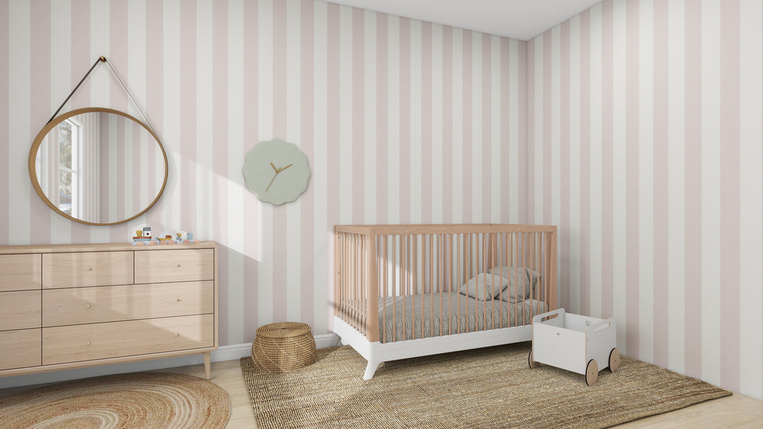 The nursery features munks and mes Circus Stripe Wallpaper Blush Rose, a wooden crib, round mirror above a light wood dresser, small white rolling toy cart, a woven mat, and a simple wall clock for added charm and quality.