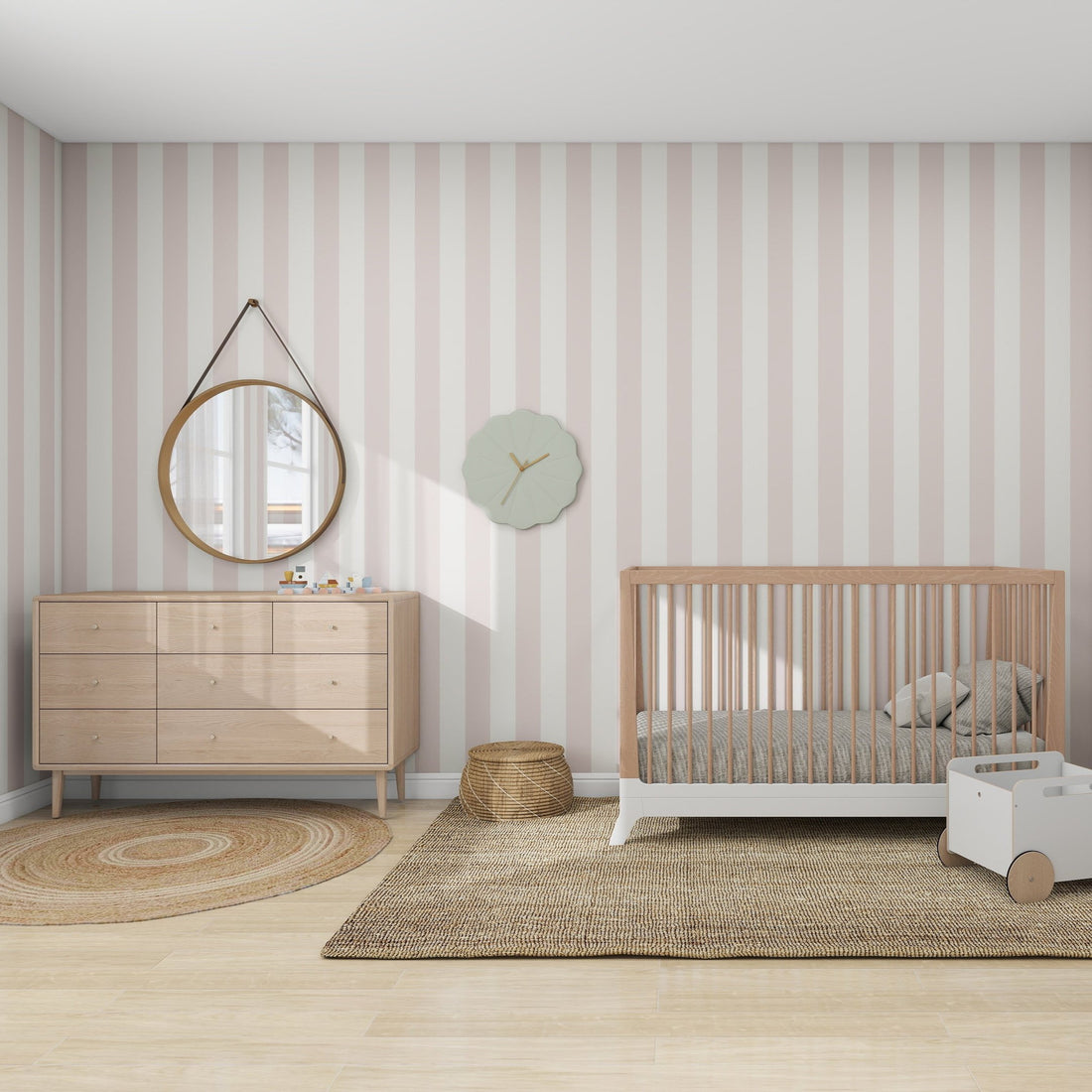 A cozy kids space with munks and mes Circus Stripe Wallpaper Blush Rose features a wooden crib, a dresser with a round mirror above, a round woven rug, small basket, decorative wall clock, white toy cart, all on an expansive beige rug.