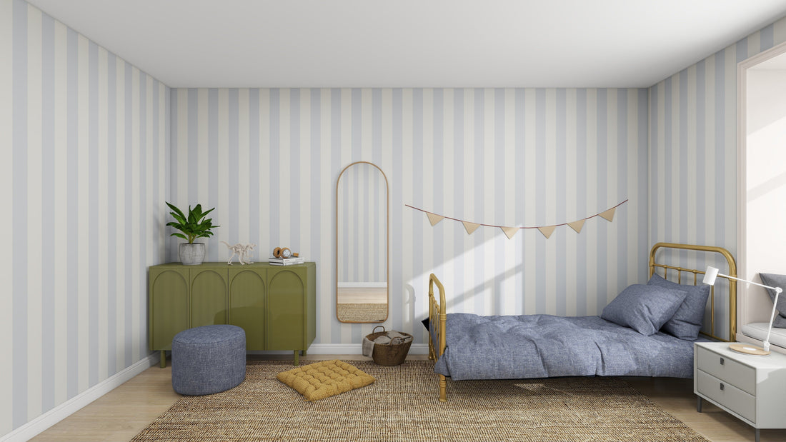 A cozy nursery features munks and mes Circus Stripe Wallpaper Sky in light blue and white. A single bed with gray bedding is next to a green dresser with plants. The wall has a tall mirror and string of pennants, while a pouf and cushion sit on the woven rug.