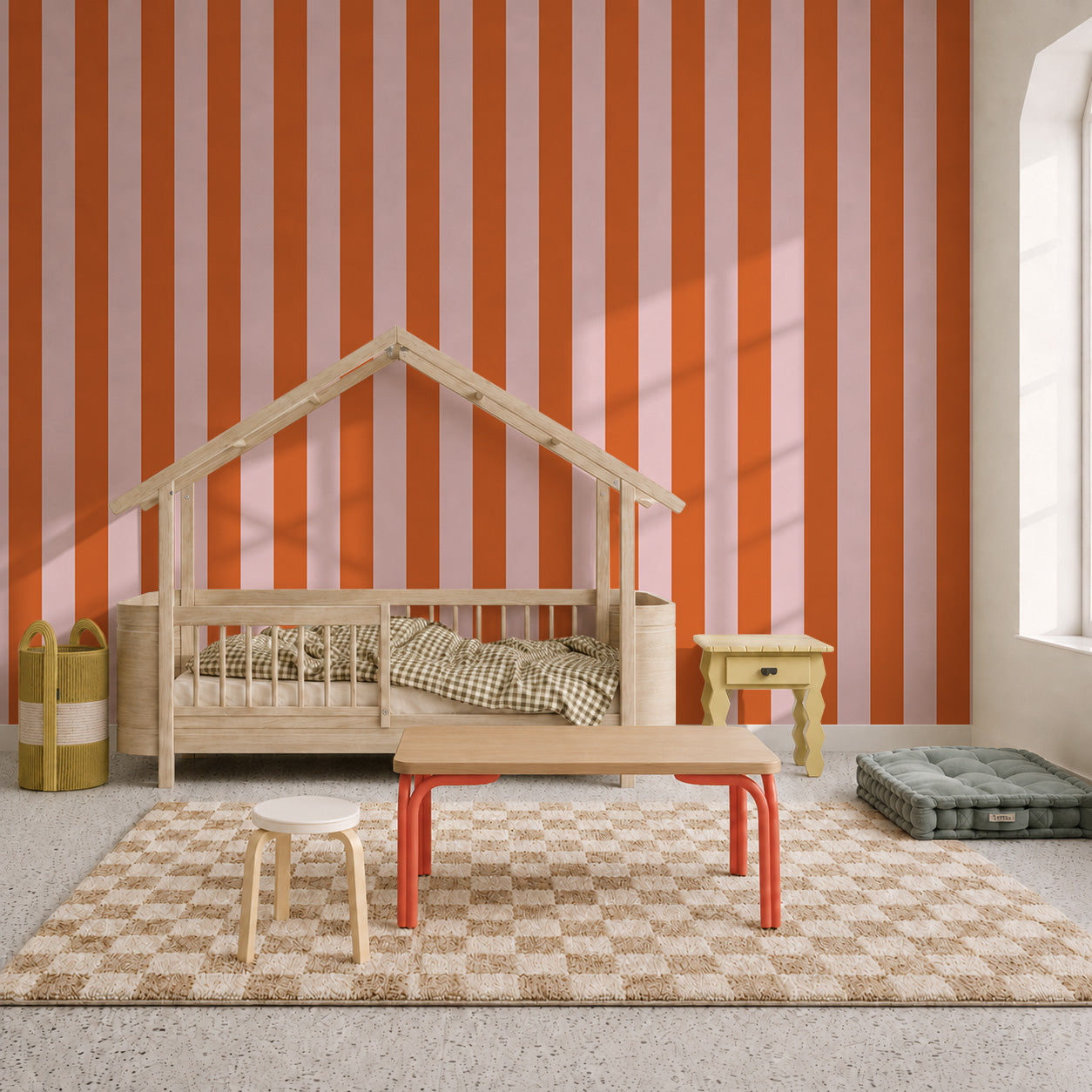 Children’s room with a bed shaped like a house, checkered rug, small tables and chairs, baskets, and a cushion. The wall is decorated with Circus Stripe Wallpaper in Pink and Red, and sunlight streams in from a window on the right.