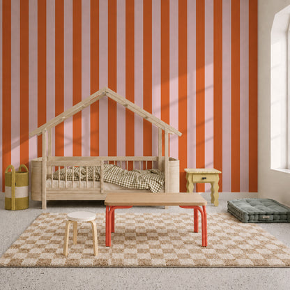 Children’s room with a bed shaped like a house, checkered rug, small tables and chairs, baskets, and a cushion. The wall is decorated with Circus Stripe Wallpaper in Pink and Red, and sunlight streams in from a window on the right.