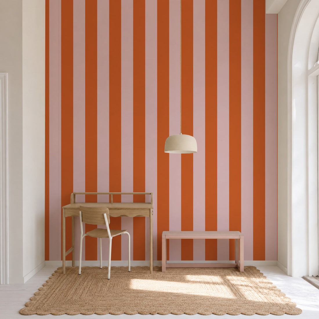 A minimalist workspace with a wooden desk and chair, a woven rug, and a pendant lamp. The wall features the Circus Stripe Wallpaper in Pink and Red, adding a playful touch to the room. A large arched window allows natural light to flood the space.
