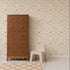 A tall brown dresser and a small beige plastic chair sit on a light-colored floor. The wall features Coles Berry Patch Wallpaper, with whimsical floral designs and pastel animal patterns that create a playful atmosphere. A beige and white checkered rug lies in front.