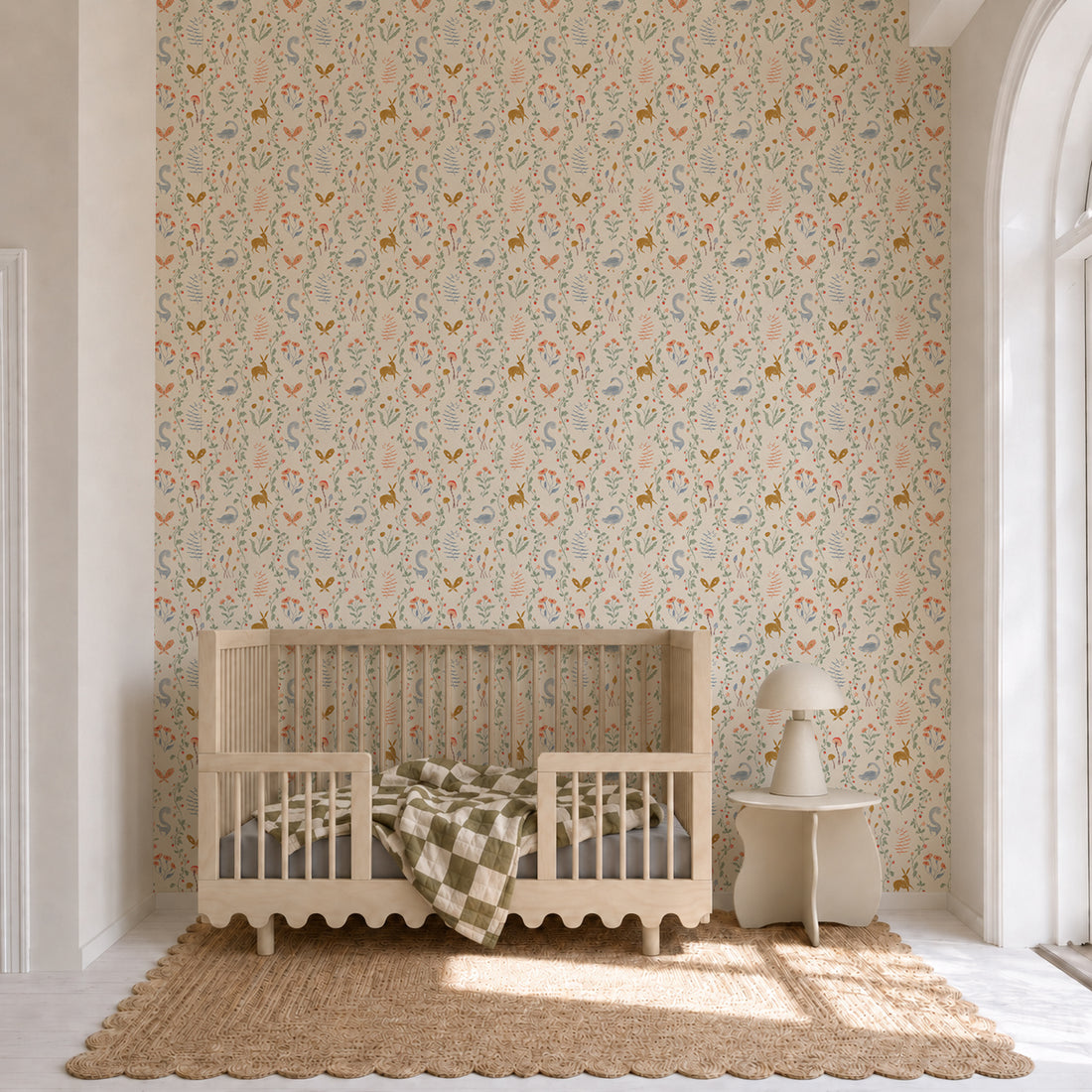 A wooden crib sits on a woven rug in a bright nursery adorned with Coles Berry Patch Wallpaper. A green and white checkered blanket hangs over the crib rail, next to a small table with a lamp by a large arched window.