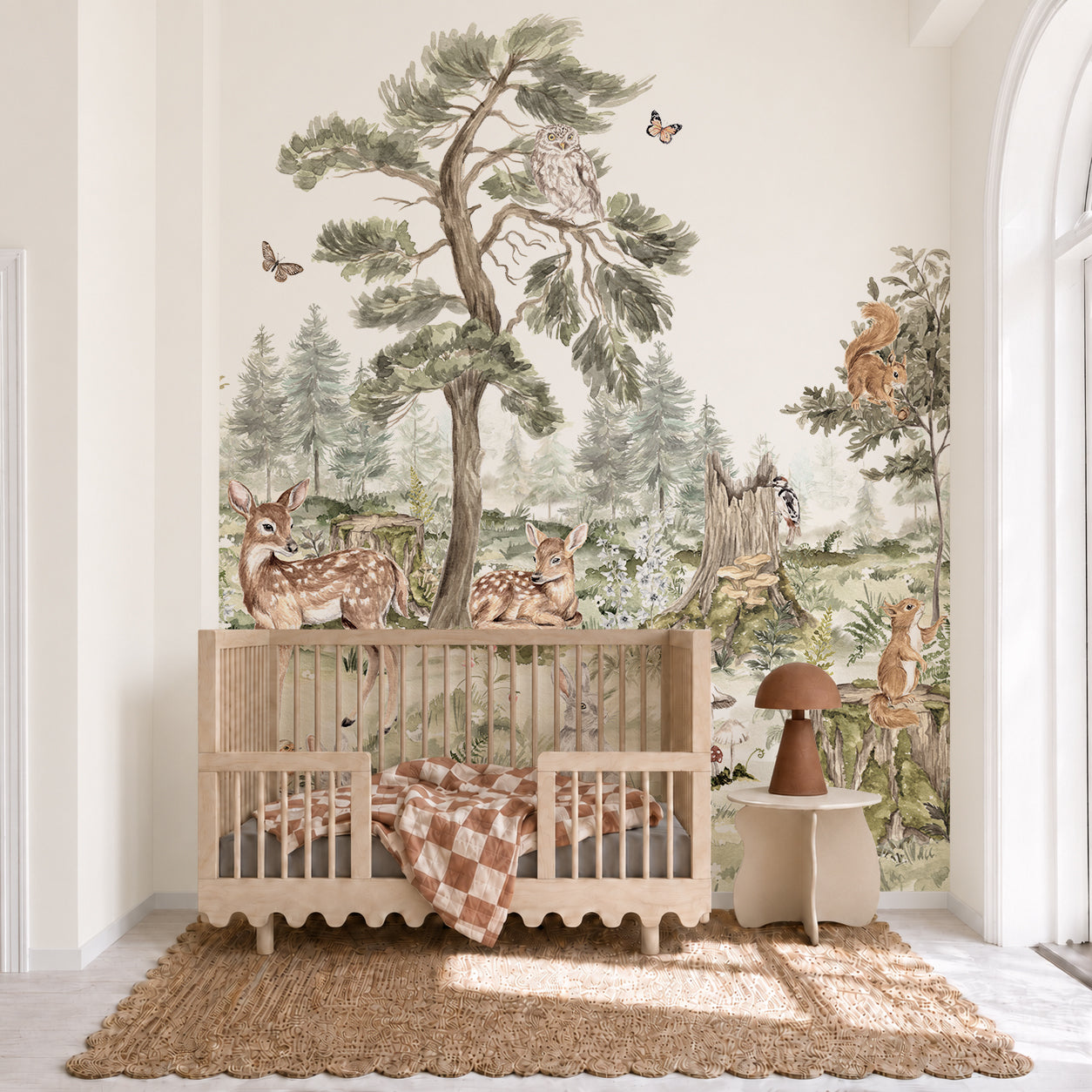 A cozy nursery features the “Into The Forest Mural,” a hand-painted woodland scene with deer, owls, squirrels, rabbits, butterflies, and trees behind a wooden crib. Natural light pours through a large arched window, illuminating the enchanting nature-inspired wall decor.