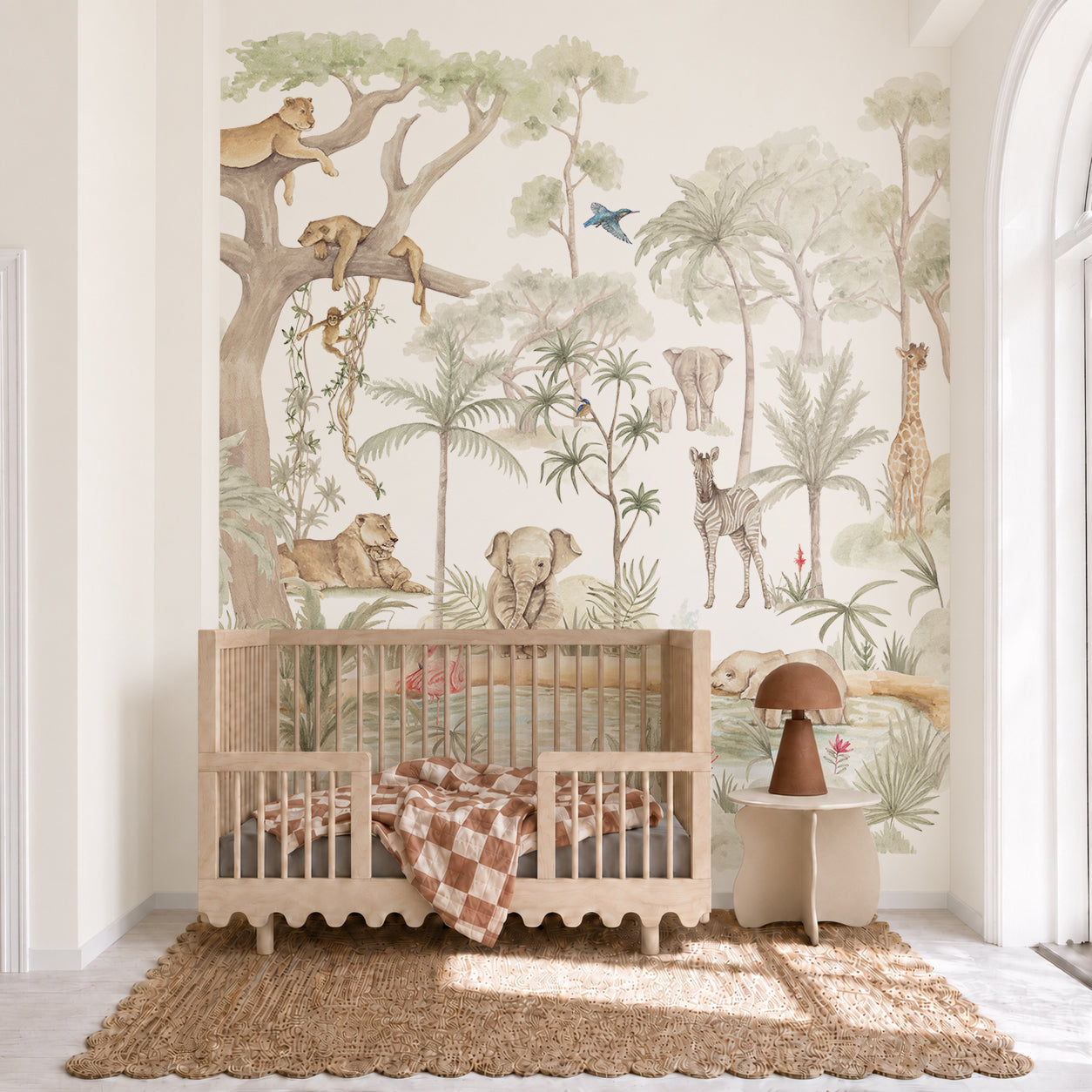 A nursery featuring a wooden crib, checkered blanket, round table, brown lamp, and a jungle-themed mural with animals such as a lion, elephant, giraffe, zebra, and monkeys amidst trees is beautifully accented with the hand-painted Safari Savannah Adventure Mural wallpaper for a whimsical touch.