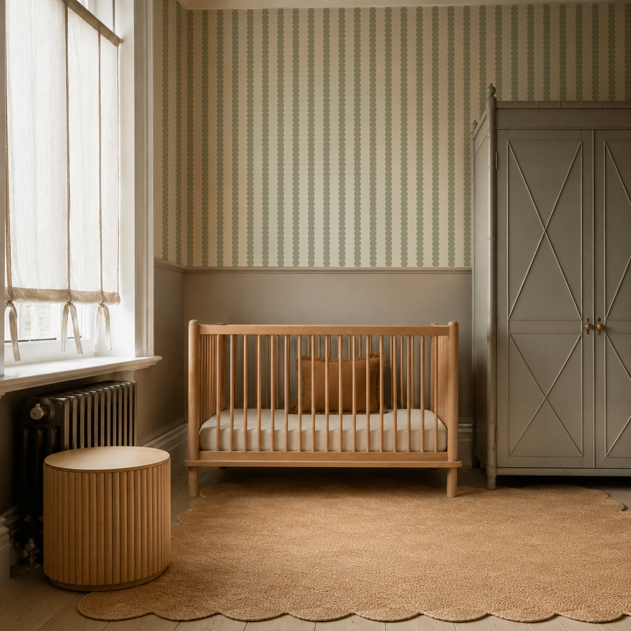 A cozy nursery with a wooden crib, a round side table, a large gray wardrobe, and a scalloped jute rug. Soft light filters through window blinds. The walls feature striped and solid paint, beautifully accented by the Scallop Stripe Wallpaper in Green for added charm.