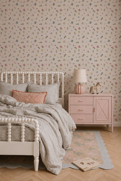 A cozy bedroom featuring a white spindle bed with gray bedding, a pink nightstand adorned with a lamp and giraffe figurines, Powdered Petals Wallpaper with hand-drawn florals, and a pastel rug scattered with books on the floor.