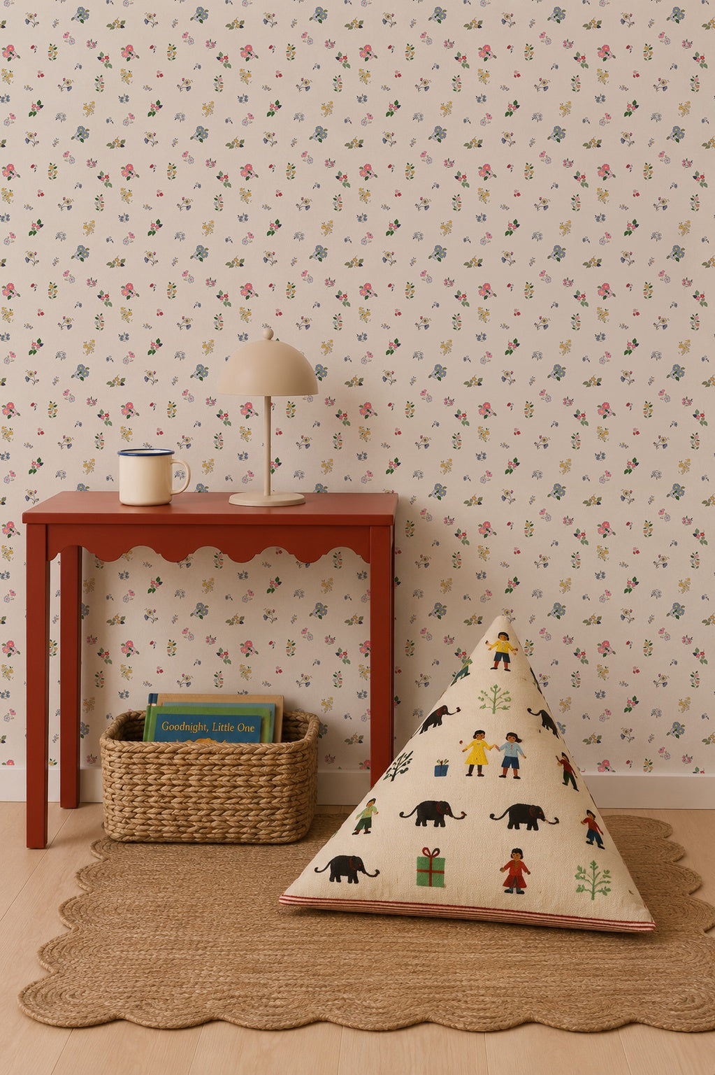 A red table with a lamp and mug stands by the Powdered Petals Wallpaper, featuring colorful, hand-drawn florals. A woven basket filled with books sits below, while a triangular cushion with animal and human prints rests on a round rug atop the wooden floor.
