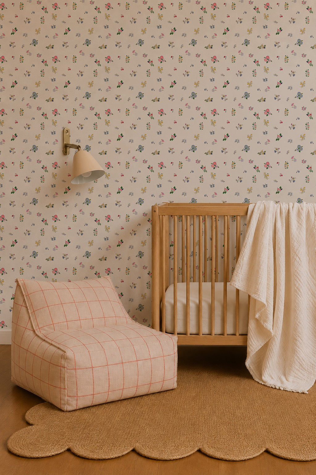 A cozy nursery featuring a wooden crib, a white textured blanket, a beige chair with a red grid pattern, a scalloped jute rug, and Powdered Petals Wallpaper. A wall-mounted lamp is positioned above the crib.