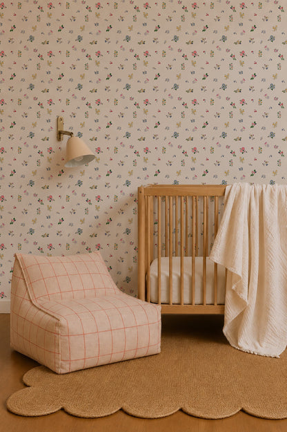 A cozy nursery featuring a wooden crib, a white textured blanket, a beige chair with a red grid pattern, a scalloped jute rug, and Powdered Petals Wallpaper. A wall-mounted lamp is positioned above the crib.