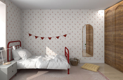 The bedroom showcases a Cherry Sprinkle Wallpaper Repeat Pattern by munks and me. A red metal bed frame with white bedding complements the red triangle pennant on the wall, with a tall mirror nearby, while a wooden wardrobe and small side table sit on a rug adorned with a plush cushion.