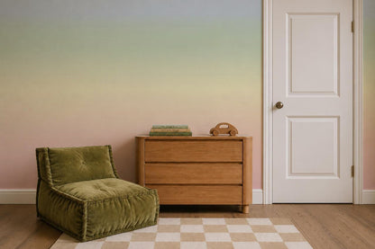 The Ombre Mural, Rainbow creates a soft pastel ombre backdrop for the calm, cozy room, which features a white door, a wooden dresser with books and a toy phone, a green cushioned chair, and a beige checkered rug on the wooden floor.