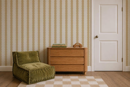 A cozy room with a green cushioned chair, a wooden dresser holding books and a rotary phone, checkered rug, light yellow Scallop Stripe Wallpaper with wavy stripes, wood floor, and a closed white door.
