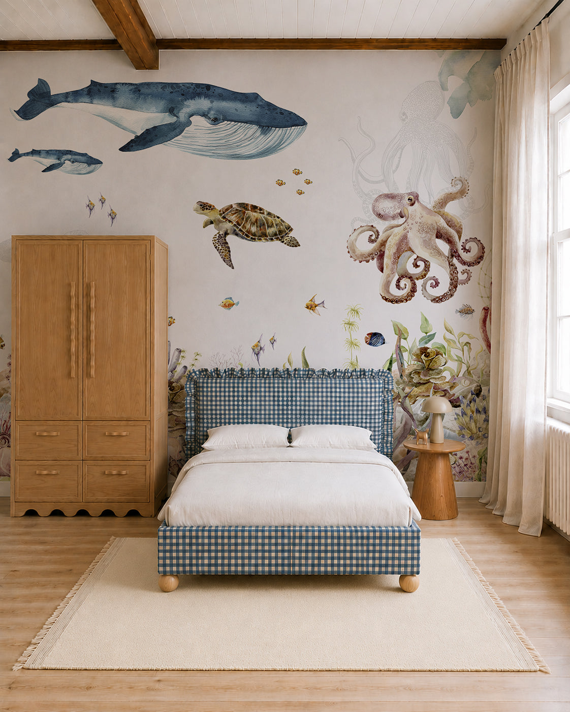 A cozy bedroom with a blue checkered bed, wooden furniture, and a cream rug features the stunning Under The Sea Mural, Watercolour wallpaper adorned with whales, an octopus, a turtle, and various sea life. Sunlight streams in through beige curtains.