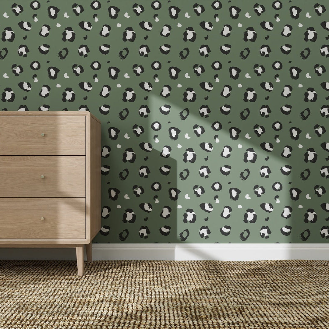 A wooden dresser stands on a woven rug against a wall featuring the Leopard Print Wallpaper Repeat Pattern in sage. Sunlight casts gentle shadows, highlighting the wallpaper’s bold yet elegant design.