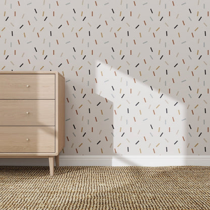 A light wood dresser sits on a woven carpet near a white wall decorated with Confetti Wallpaper Repeat Pattern. Sunlight casts window-shaped shadows, highlighting the wallpapers lively, playful design.