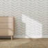 A light wood dresser sits on a woven tan carpet against a wall covered with Herringbone Wallpaper Repeat Pattern | Blue, adding a modern touch. Sunlight casts shadows, creating a bright and airy atmosphere.