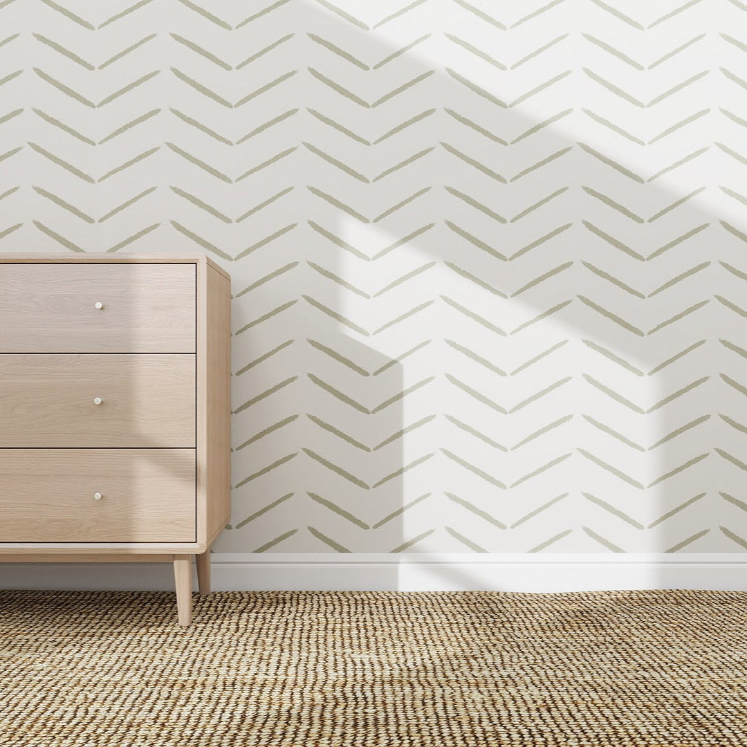 A light wooden dresser stands on a woven rug beside a white wall decorated with Herringbone Wallpaper Repeat Pattern | Olive. Sunlight casts sharp shadows, adding a stylish touch to the serene setting.