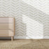 A light wooden dresser stands on a woven rug beside a white wall decorated with Herringbone Wallpaper Repeat Pattern | Olive. Sunlight casts sharp shadows, adding a stylish touch to the serene setting.
