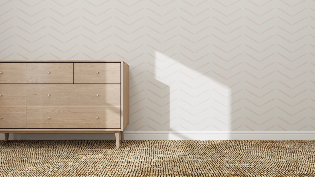 A light wood dresser with six drawers sits on a woven rug against a white wall decorated with Herringbone Wallpaper Repeat Pattern | Rose, while sunlight casts geometric shadows that enhance the calm atmosphere of the space.