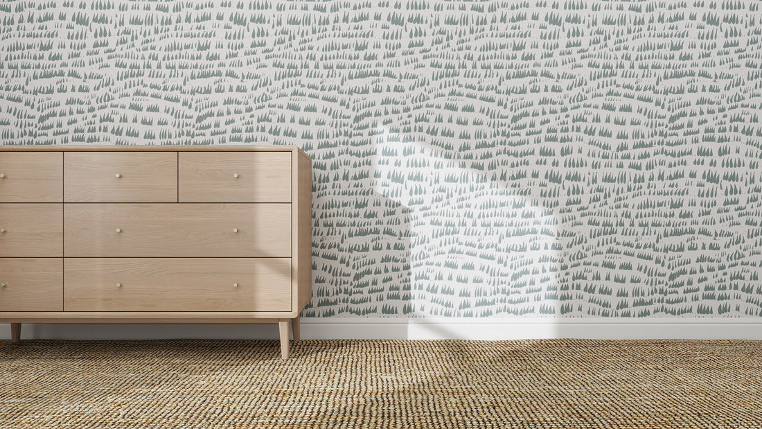 A light wooden dresser stands on a woven rug against a wall decorated with Daphne Drops Wallpaper Repeat Pattern | Blue, its abstract raindrop motif softly highlighted by sunlight casting gentle shadows in the room.