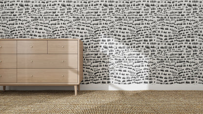 A light wooden dresser stands on a woven rug beside a wall adorned with Daphne Drops Wallpaper Repeat Pattern | Charcoal. Sunlight streams across the floor and part of the wall.
