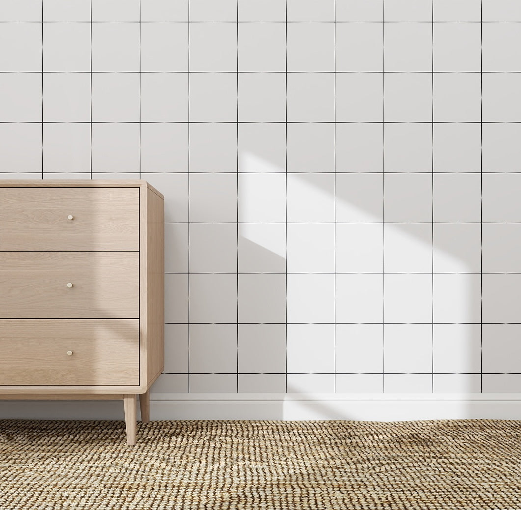 A light wood dresser with three drawers stands on a woven rug by a white tiled wall featuring the Urban Grid Wallpaper Repeat Pattern, creating a contemporary vibe as sunlight casts shadows across the wall and floor.